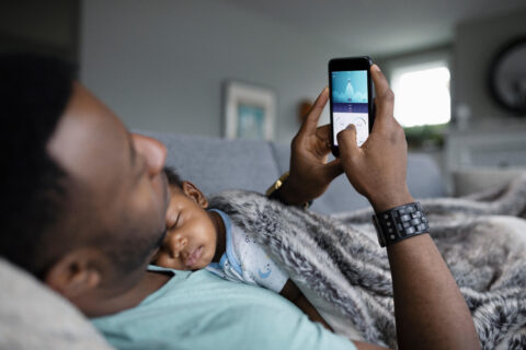 Father using smart phone with baby son sleeping on chest